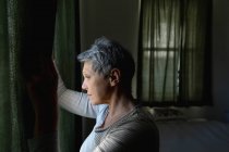 Nahaufnahme einer reifen kaukasischen Frau mit kurzen grauen Haaren, die die Vorhänge zieht und zu Hause aus dem Fenster schaut — Stockfoto