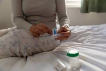 Вид спереди средней части женщины, сидящей на своей кровати дома, принимающей лекарства из еженедельной коробки для таблеток, с другими контейнерами лекарств на кровати рядом с ней — стоковое фото