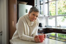 Portrait d'une femme blanche mature aux cheveux gris courts assise dans sa cuisine tenant une tasse de café et regardant la caméra sourire — Photo de stock