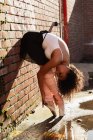 Vista lateral de cerca de una joven bailarina de ballet de raza mixta de pie sobre sus dedos contra una pared de ladrillo, doblada doble sosteniendo sus talones, con la cabeza vuelta, en la azotea de un edificio urbano - foto de stock