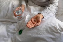 Вид спереду середня секція жінки, що сидить у ліжку вдома, тримає таблетку і склянку води, пляшку таблеток на ліжку поруч з нею — стокове фото