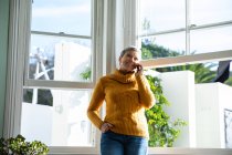 Vorderansicht einer reifen kaukasischen Frau, die vor den Fenstern ihres Wohnzimmers steht und mit einem Smartphone spricht — Stockfoto