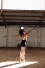 Боковой вид молодой танцовщицы балета смешанной расы в пуантах, стоящей на пальцах ног в лучах солнца с протянутыми руками во время танцев в пустой комнате на заброшенном складе — стоковое фото