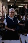 Portrait rapproché d'une femme de race mixte d'âge moyen debout à une table tenant un chapeau dans l'atelier d'une usine de chapeaux, entourée d'équipements et de matériaux — Photo de stock