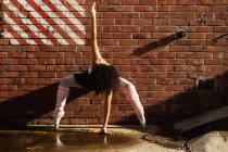 Vue de face d'une jeune danseuse de ballet mixte debout sur ses orteils contre un mur de briques avec un bras levé et une main touchant le sol, sur le toit d'un bâtiment urbain — Photo de stock