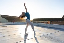 Vista laterale di una giovane ballerina di danza mista che si appoggia indietro in una posa di balletto con le braccia tese, sul tetto di un edificio urbano, retroilluminata dalla luce del sole — Foto stock