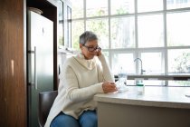 Side view of a mature Caucasian woman with short grey hair wearing glasses sitting in her kitchen reading about her medication, with pill bottles, a weekly pill box and a glass of water on the counter beside her — Stock Photo