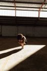 Вид спереди молодой танцовщицы балета смешанной расы в пуантах, приседающей на коленях, балансирующей на пальцах ног в лучах солнца, танцующей в пустой комнате на заброшенном складе — стоковое фото