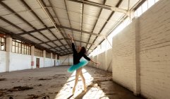 Боковой вид молодой танцовщицы смешанной расы в синей пачке и пуантах, танцующей на пальцах ног в лучах солнечного света в пустой комнате на заброшенном складе — стоковое фото
