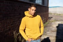 Vista frontal de perto de um jovem vestindo um capuz amarelo de pé ao sol com as mãos nos bolsos do lado de fora de um armazém abandonado — Fotografia de Stock