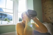 Vista lateral de cerca de una mujer caucásica madura con el pelo gris corto sentada en casa en su sala de estar con un auricular VR, iluminada por la luz del sol desde una ventana detrás de ella - foto de stock