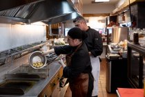 Vista lateral de perto de um chefe masculino caucasiano de meia idade supervisionando o trabalho de uma jovem chef feminina caucasiana, drenando batatas em uma cozinha do restaurante — Fotografia de Stock