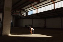 Вид сбоку на молодую танцовщицу балета смешанной расы в пуантах, приседающую на ногах, сбалансированную на солнечных лучах, танцующую в пустой комнате на заброшенном складе — стоковое фото