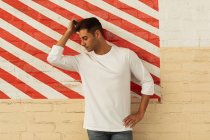 Front view of a young man leaning against a wall scratching his head, which is turned to the side, and with is hand on his hip in an empty room with a section of red and white striped wall at an abandoned warehouse — Stock Photo