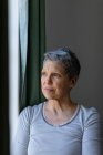Front view close up of a mature Caucasian woman with short grey hair standing and looking out of the window at home — Stock Photo