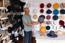 Side view of a senior mixed race man standing and inspecting a finished hat, surrounded by hats on display in the showroom at a hat factory — Stock Photo
