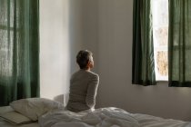 Rear view of a mature Caucasian woman with short grey hair sitting on the side of her bed at home looking out of the window — Stock Photo
