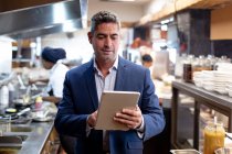 Front view close up of a middle aged Caucasian male restaurant manager using a tablet computer in a busy restaurant kitchen, while kitchen staff work in the background — Stock Photo
