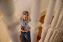 Vista frontal elevada de una mujer caucásica madura con pelo gris corto sentada en sus escaleras en casa usando un teléfono inteligente - foto de stock