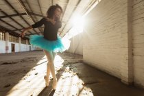 Nahaufnahme einer jungen Balletttänzerin mit gemischter Rasse, die ein blaues Tutu und Spitzenschuhe trägt und im Sonnenlicht in einem leeren Raum einer verlassenen Lagerhalle auf ihren Zehen tanzt — Stockfoto