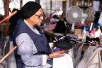 Vista lateral de perto de uma mulher de meia-idade mista segurando e inspecionando um pequeno chapéu na oficina em uma fábrica de chapéus, cercada por materiais — Fotografia de Stock