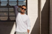 Ritratto di un giovane uomo che indossa occhiali da sole in piedi contro un muro in un pozzo di luce solare con una mano dietro la testa, guardando la telecamera in un magazzino abbandonato — Foto stock
