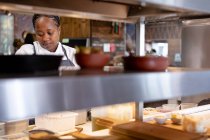 Front view close up of a young African American female chef working in a restaurant kitchen, seen through shelves — Stock Photo