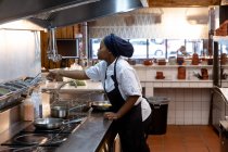 Вид сбоку на молодую афроамериканку-повара, которая тянется за ингредиентами на кулинарной станции на кухне ресторана — стоковое фото
