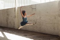 Seitenansicht einer jungen Balletttänzerin mit gemischter Rasse, die mit erhobenen Armen in die Luft springt, während sie in einem leeren Raum einer verlassenen Lagerhalle tanzt — Stockfoto