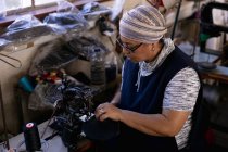 Вид збоку крупним планом жінка середнього віку змішаної раси, що використовує швейну машинку на капелюшній фабриці, оточена обладнанням та матеріалами — стокове фото