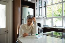 Vista frontal de una mujer caucásica madura con pelo gris corto sentada en su cocina mirando su medicación, con frascos de pastillas, una caja de pastillas semanal y un vaso de agua en el mostrador junto a ella - foto de stock