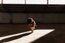Vue de face d'une jeune danseuse de ballet mixte portant des chaussures à pointe accroupie tenant ses genoux équilibrés sur ses orteils dans un rayon de soleil tout en dansant dans une pièce vide dans un entrepôt abandonné — Photo de stock