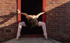 Вид спереди на молодую балетную танцовщицу смешанной расы, держащую танцевальную позу и смотрящую вниз в дверях в кирпичной стене на крыше городского здания — стоковое фото