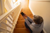 Підвищений задній вигляд зрілої кавказької жінки з коротким сивим волоссям, сидячи на сходах будинку, використовуючи смартфон, з сонячним світлом у фоновому режимі — стокове фото
