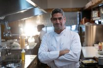 Retrato close-up de um sorriso de meia-idade caucasiano chef masculino vestindo brancos de pé com os braços cruzados em uma cozinha restaurante — Fotografia de Stock