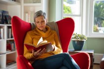 Вид спереди на взрослую белую женщину с короткими седыми волосами, сидящую в красном кресле в гостиной и читающую книгу, с окном на заднем плане — стоковое фото