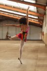 Rückansicht einer jungen Balletttänzerin mit gemischter Rasse, die mit ausgestreckten Armen auf einem Bein steht, während sie in einem leeren Raum einer verlassenen Lagerhalle tanzt — Stockfoto