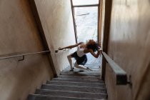 Vista lateral elevada de uma jovem dançarina de balé mista segurando os corrimãos e dançando no fundo de uma escada em um armazém abandonado — Fotografia de Stock