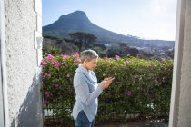 Die Seitenansicht einer reifen kaukasischen Frau mit kurzen grauen Haaren, die mit einem Smartphone in ihrem Garten steht, ist von der Haustür aus zu sehen, mit einer ländlichen Szene und einem Berg im Hintergrund. — Stockfoto