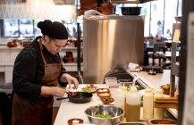 Вид збоку крупним планом молодого Кавказького жіночого кухаря додавання інгредієнтів на сковороду в ресторані кухні — стокове фото