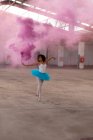 Frontansicht einer jungen Balletttänzerin mit gemischter Rasse, die ein blaues Tutu und Spitzenschuhe trägt und in einem leeren Raum einer verlassenen Lagerhalle mit einer rosa Rauchgranate tanzt — Stockfoto