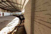 Seitenansicht eines jungen männlichen Balletttänzers, der in einem leeren Raum einer verlassenen Lagerhalle in Schwarz gegen eine Wand tanzt — Stockfoto