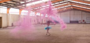 Vue arrière d'une jeune danseuse de ballet mixte portant un tutu bleu et des chaussures pointes dansant tenant une grenade de fumée rose dans une pièce vide dans un entrepôt abandonné — Photo de stock