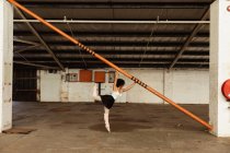 Side view of a young mixed race female ballet dancer wearing pointe shoes dancing standing on one leg on her toes holding a structural pole in an empty room at an abandoned warehouse — Stock Photo