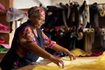 Vista laterale da vicino di una donna di razza mista di mezza età in piedi a un tavolo che lavora con tessuto giallo in una fabbrica di cappelli . — Foto stock