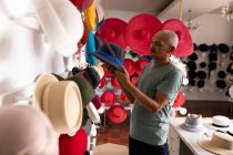 Side view of a senior mixed race man standing and inspecting a finished hat, surrounded by hats on display in the showroom at a hat factory — Stock Photo