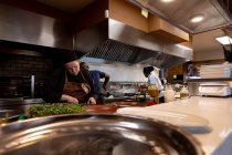 Vue de face d'une jeune cuisinière caucasienne hachant des ingrédients à un comptoir dans une cuisine de restaurant occupée, vue sur un comptoir et derrière un équipement de cuisine, avec d'autres employés de cuisine travaillant en arrière-plan — Photo de stock