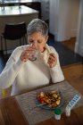Elevated front view of a mature Caucasian woman sitting at her dining table drinking a glass of water, with food on a plate and bottles of tablets and pills on the table in front of her — Stock Photo
