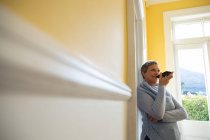 Боковой вид взрослой белой женщины с короткими седыми волосами, прислонившейся к стене дома, разговаривающей на смартфоне, который она держит перед своим ртом и улыбающейся, с сельской сценой, видимой через окно на заднем плане — стоковое фото