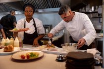 Front view close up of a middle aged Caucasian male chef plating up dishes while a young African American female chef holds a prepared dish for serving in a busy restaurant kitchen, with staff working the background — Stock Photo
